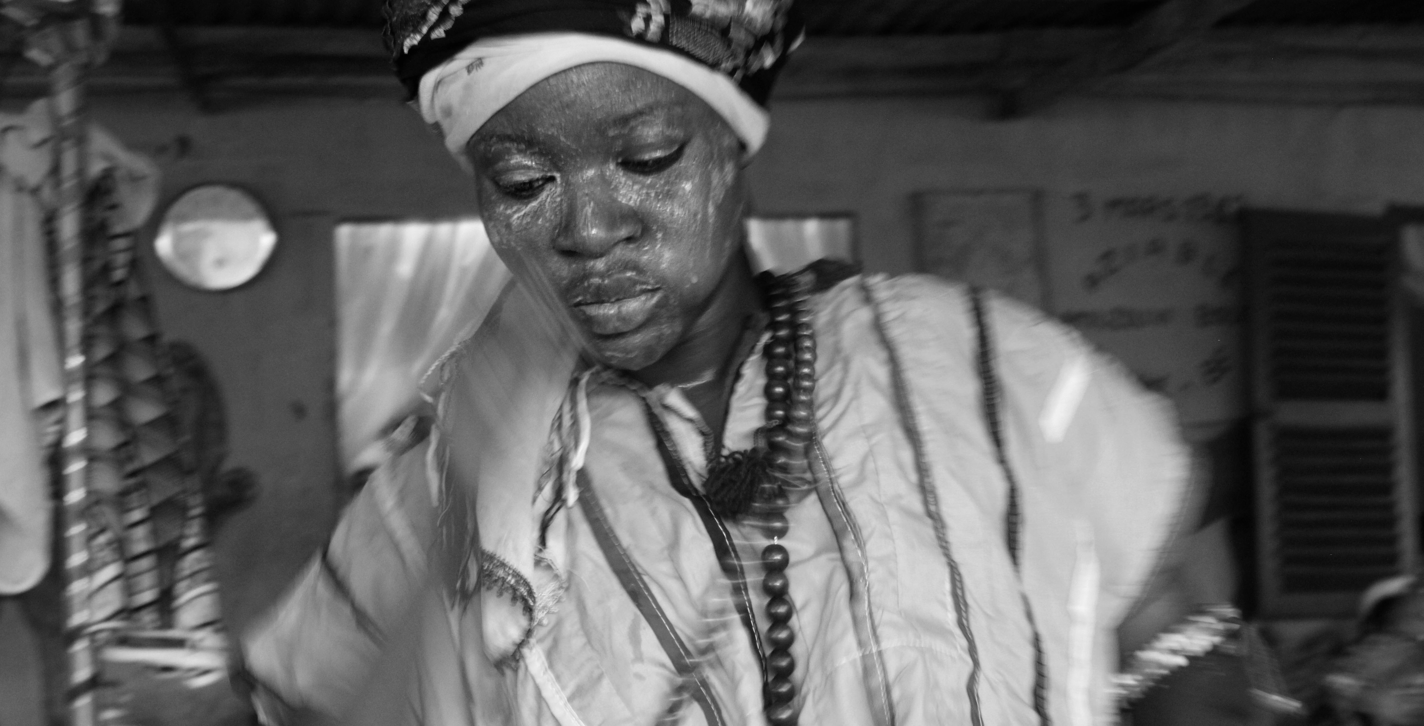 A black and white image of a black woman wearing traditional headress and beads. She also has what appears to be white paint on her face.